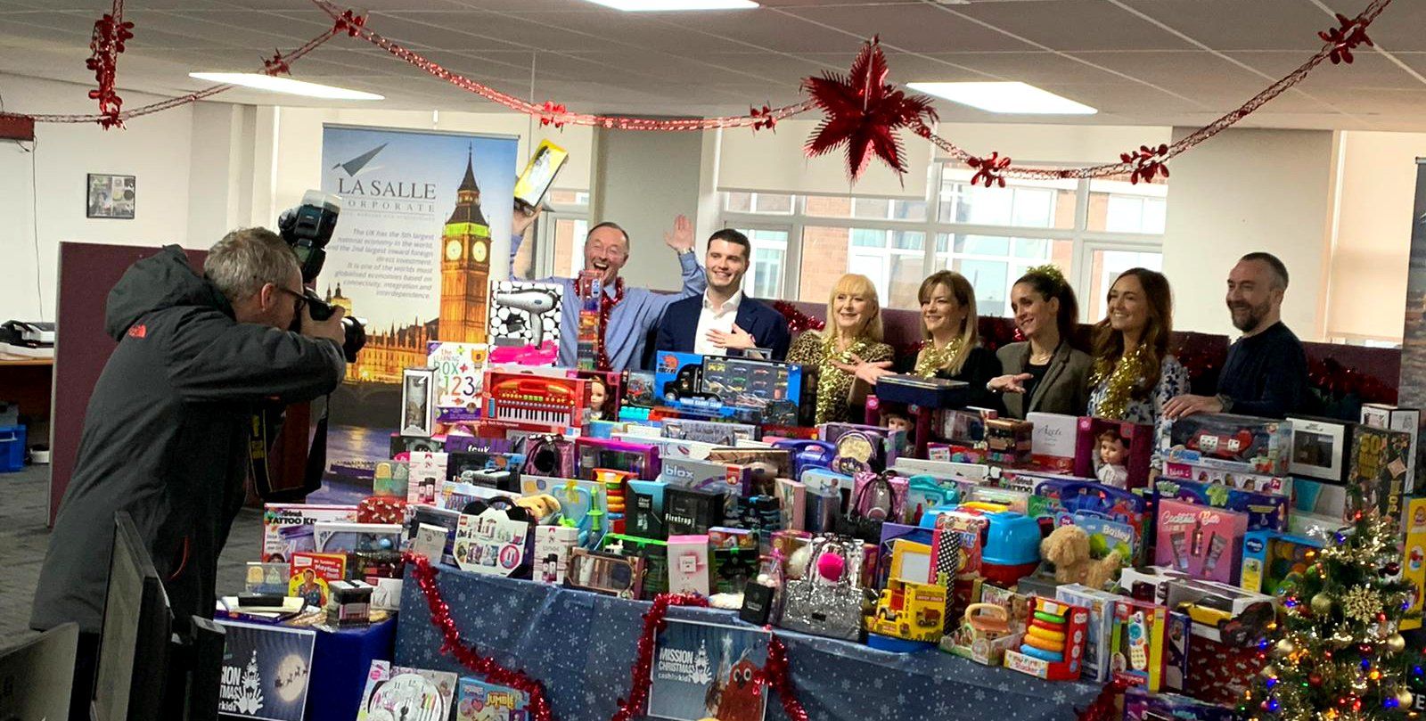 Volunteers behind a table stacked with colorful donated toys at a festive indoor holiday drive