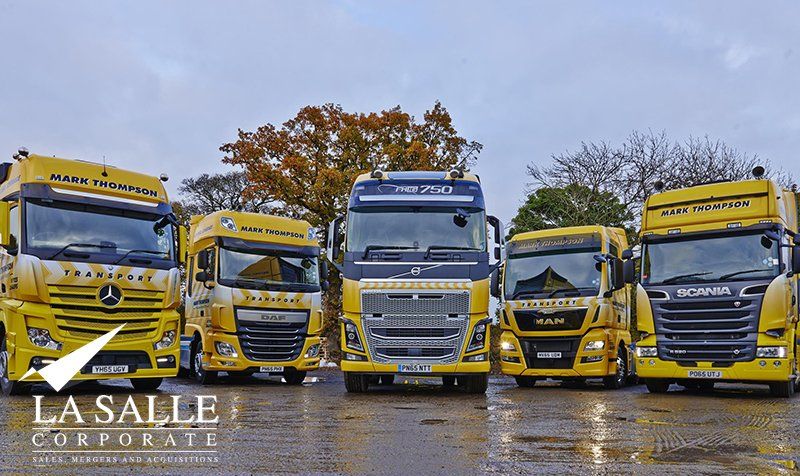 Five yellow and white trucks lined up on wet pavement with autumn trees in the background