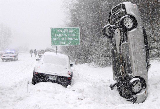 Snowy roadside with an overturned car beside a buried white car and a road sign during a blizzard