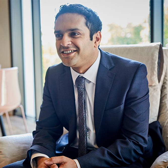 Smiling man in a navy suit seated in a bright office near a window.