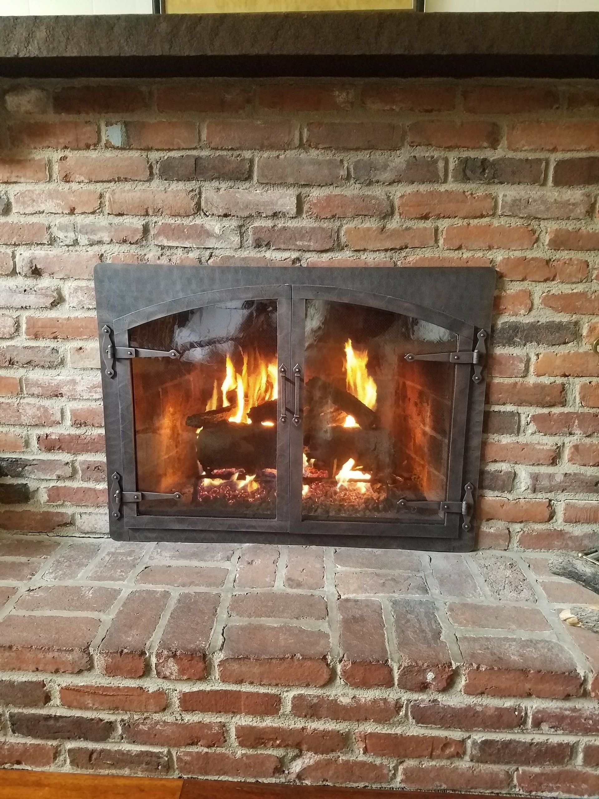 Fireplace with roaring fire, enclosed in glass doors against a red brick wall.