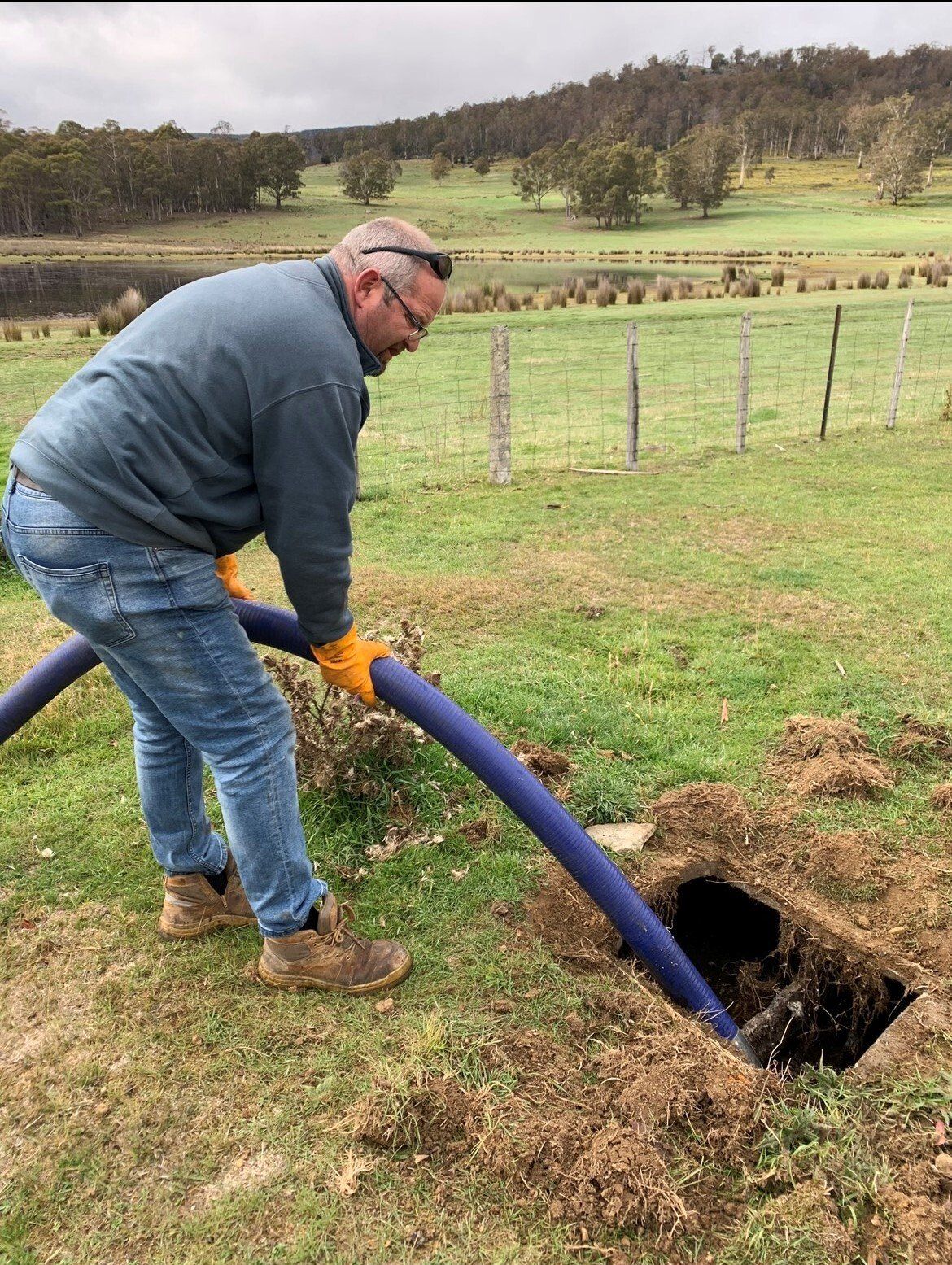 Man pumping septic tank with blue hose in a grassy field.