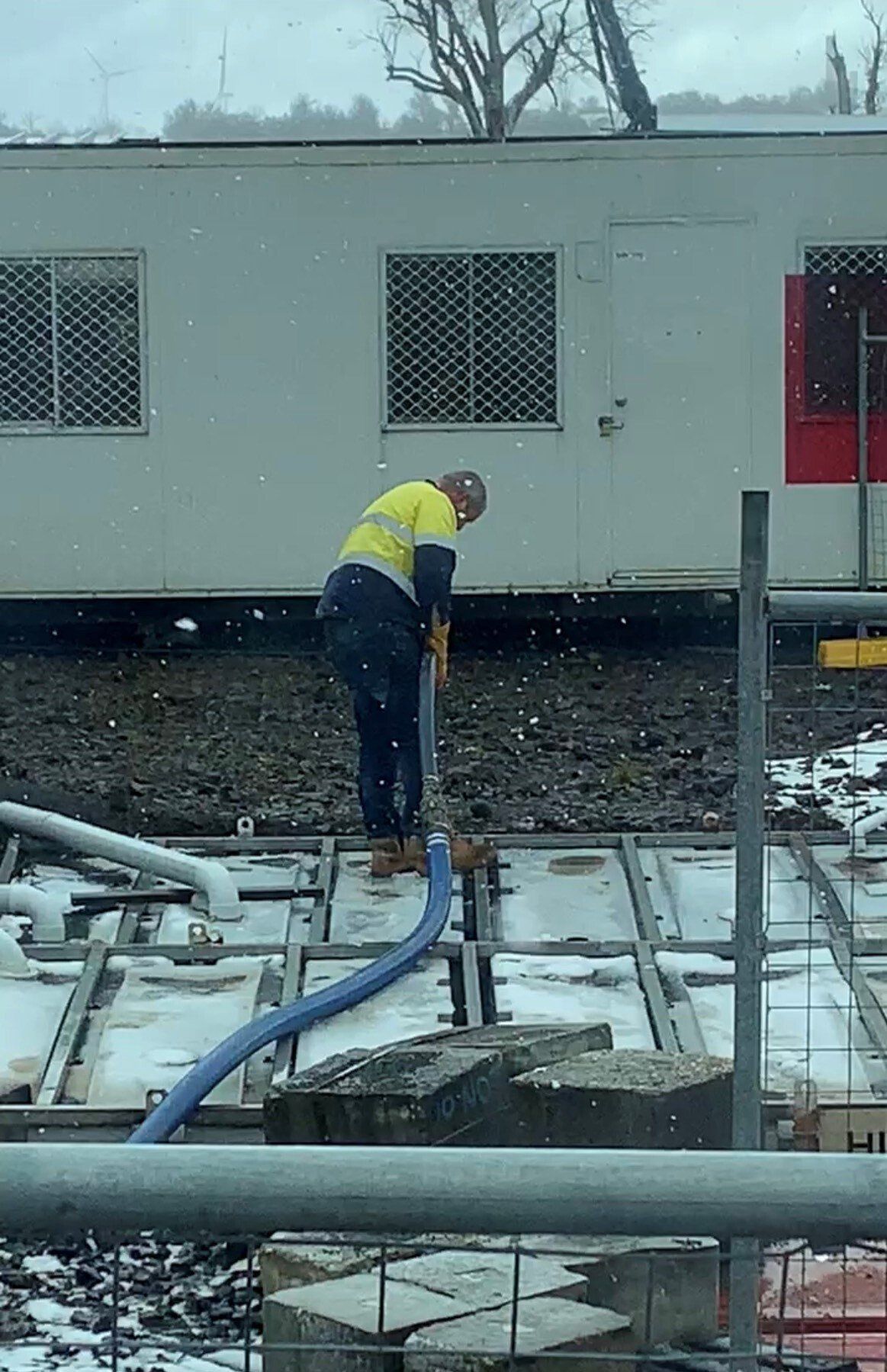Construction worker in yellow vest connecting a blue hose to a metal grid in a snowy setting.