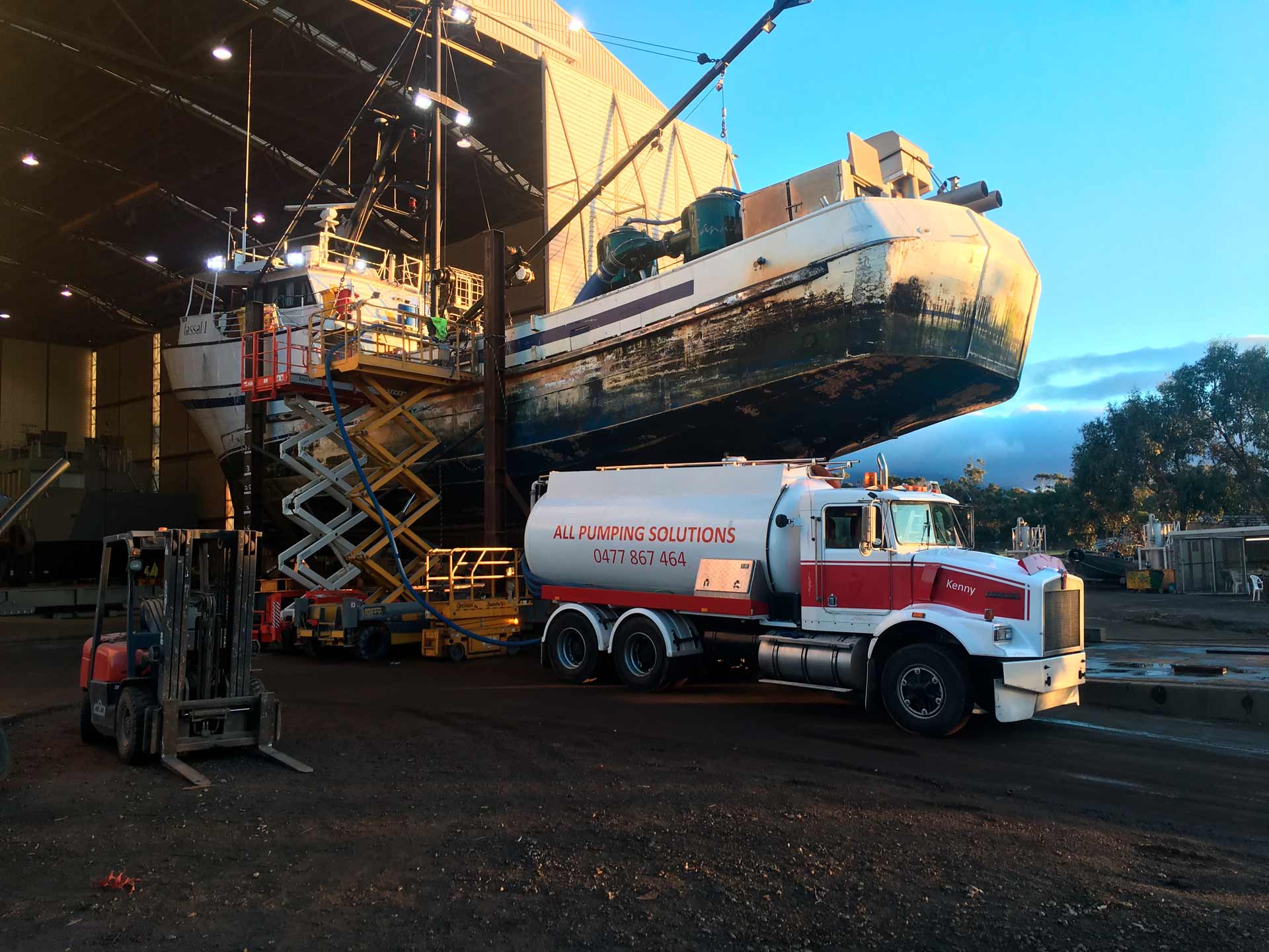 A ship being lifted by a crane above a red and white tanker truck at a shipyard.
