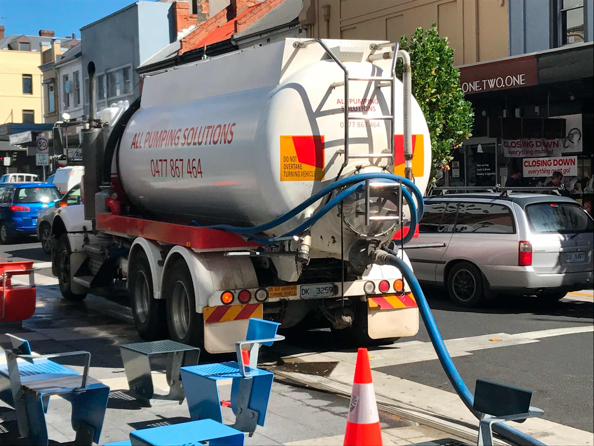 Tanker truck parked on a street, connected by a blue hose to a drain.