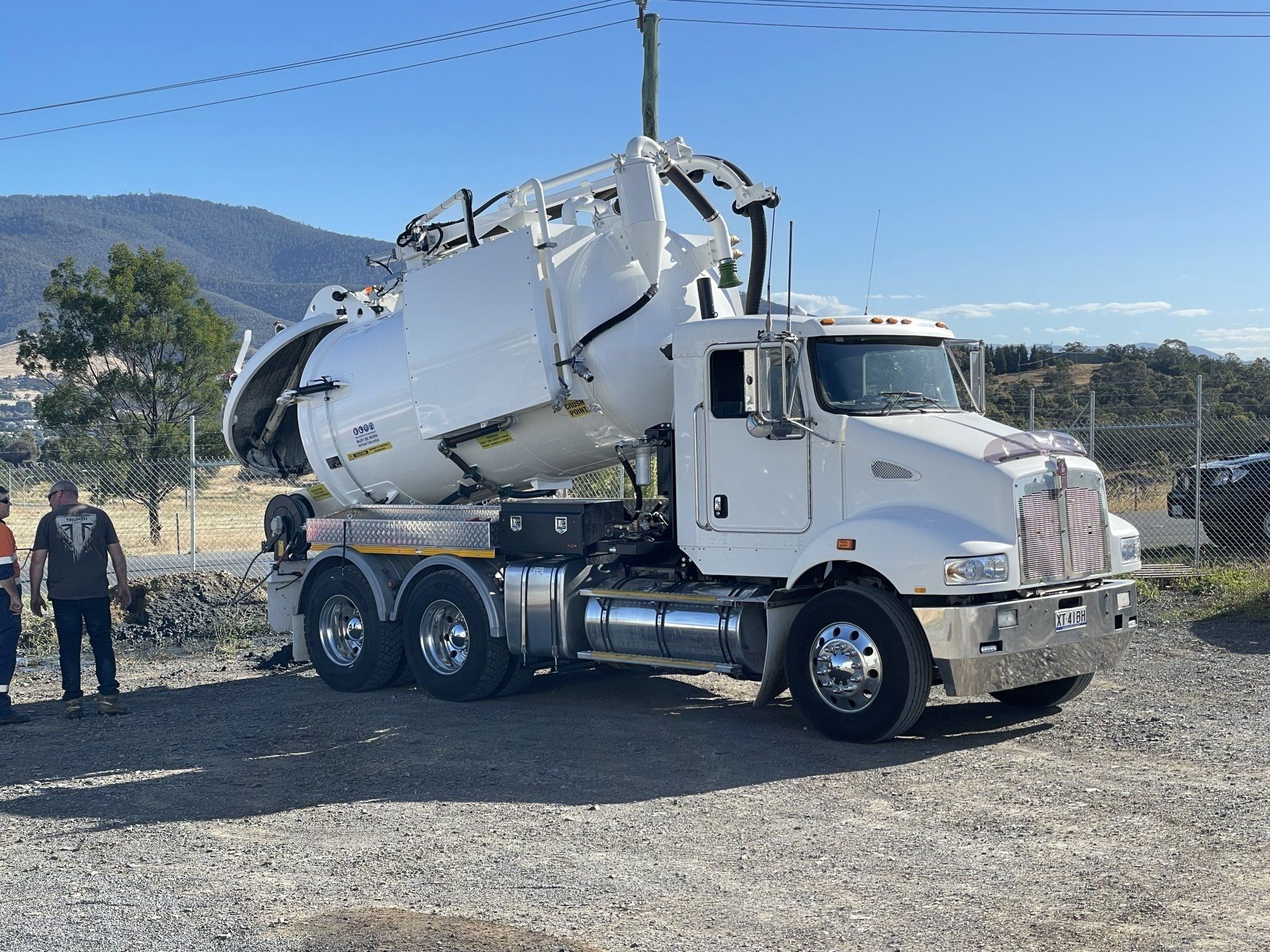 White vacuum truck with a raised tank, parked outdoors with people and hills in the background.