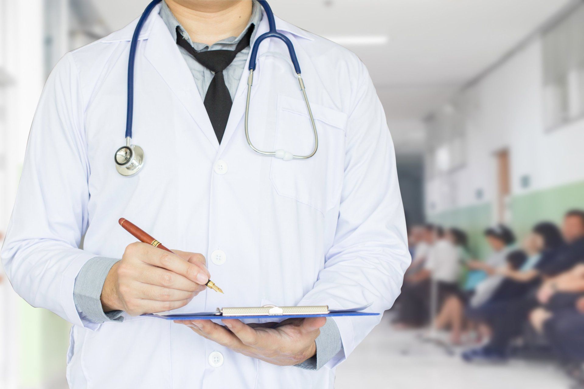 A doctor with a stethoscope around his neck is holding a clipboard and writing on it.
