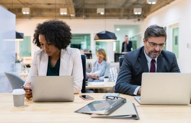 A man and a woman are sitting at a desk with laptops.