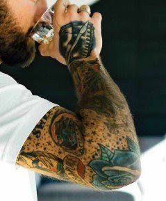 A man with a lot of tattoos on his arms is drinking from a glass