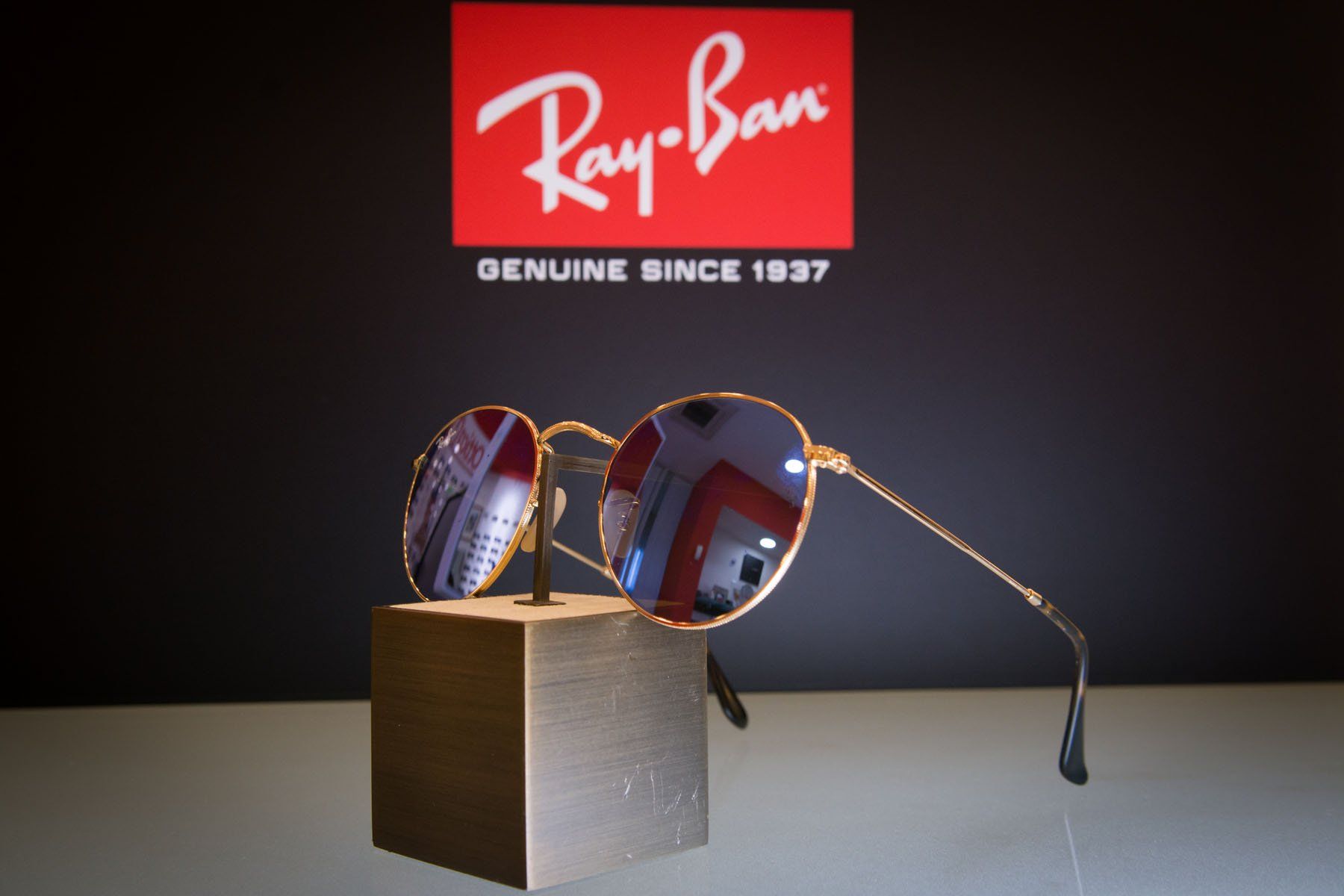Gli occhiali da sole Ray-Ban con montatura dorata sono appoggiati su un piccolo blocco, su uno sfondo nero e sul logo Ray-Ban.