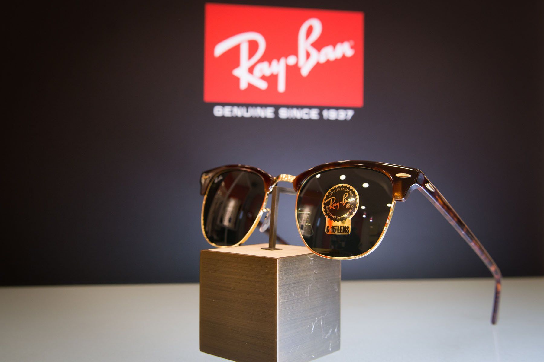 Occhiali da sole Ray-Ban esposti su un espositore con logo sullo sfondo; montatura marrone e oro.