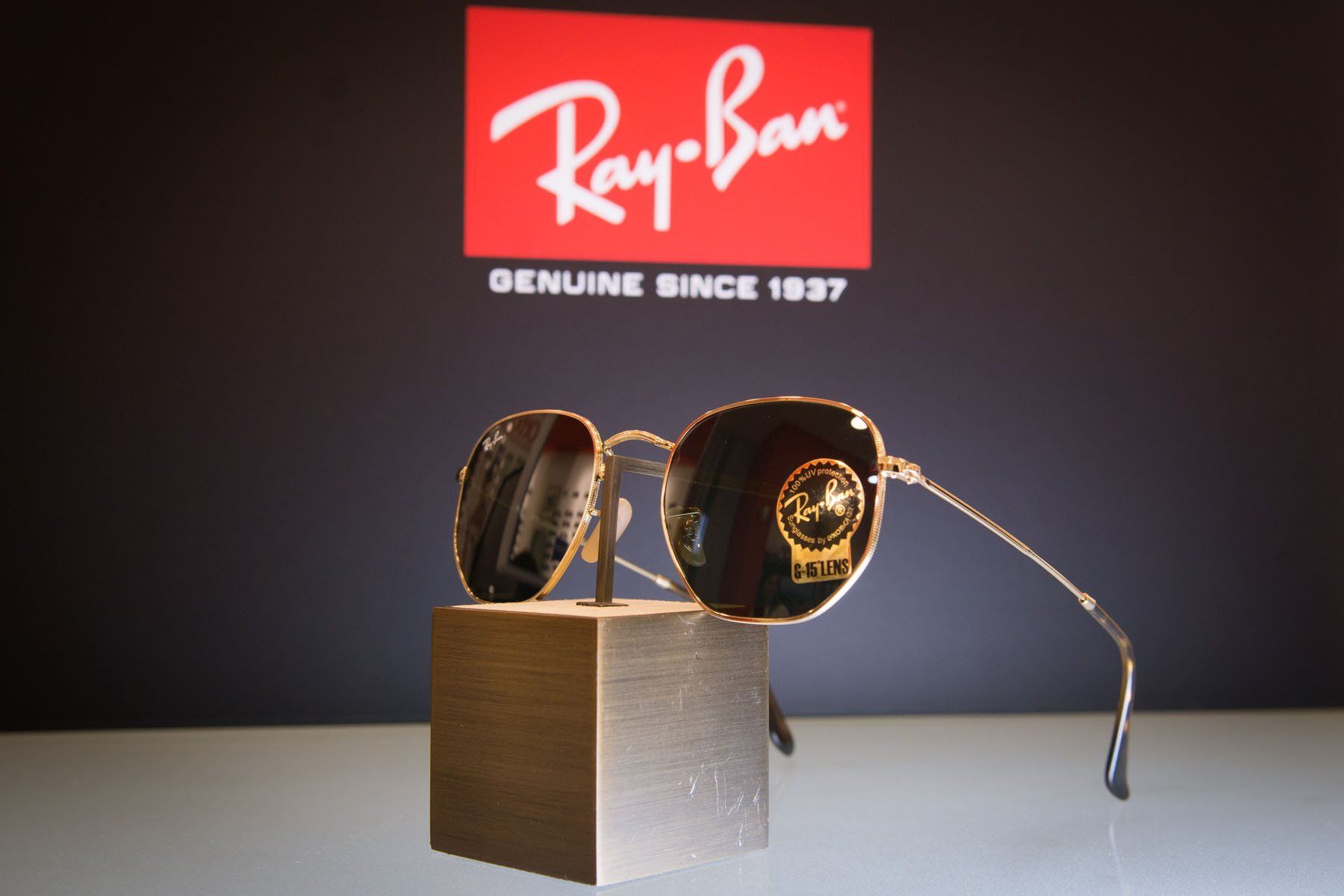 Occhiali da sole Ray-Ban su un supporto dorato, davanti al logo Ray-Ban su uno sfondo nero.