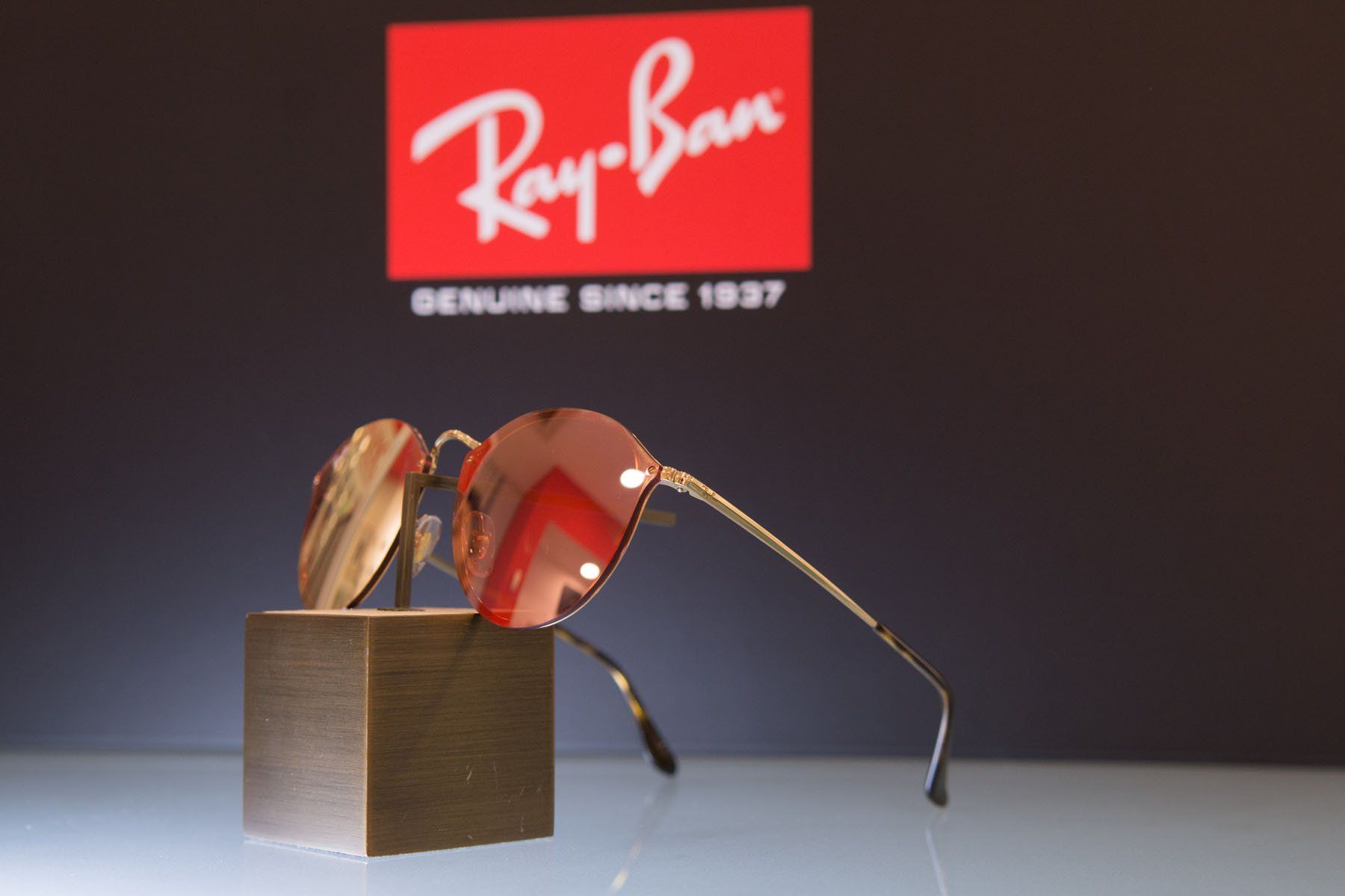 Occhiali da sole Ray-Ban esposti su un espositore, davanti al logo Ray-Ban. Montatura dorata, lenti rosa.