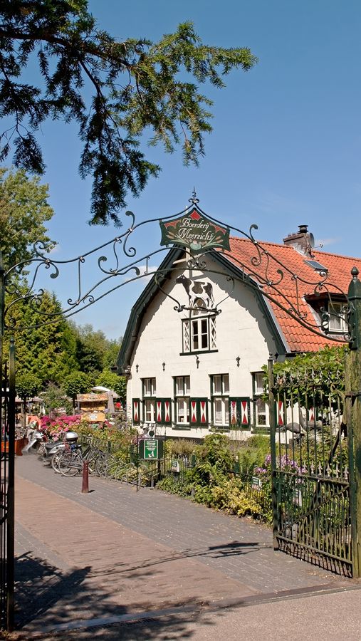 Een sierlijke ijzeren poort leidt naar een wit gebouw met een rood pannendak, een tuin en bomen.