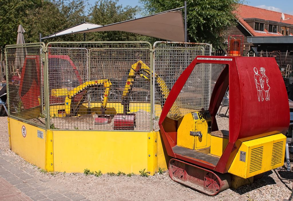 Een geel en rood speelgoedbouwvoertuig voor kinderen, met daarin verschillende speelgoedgraafmachines, in een omheinde ruimte.