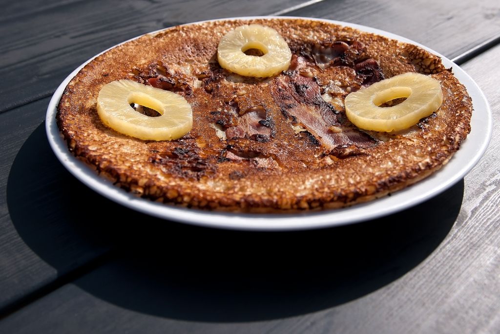 Een pannenkoek met ananasringen op een wit bord, geplaatst op een donkere houten tafel in het zonlicht.