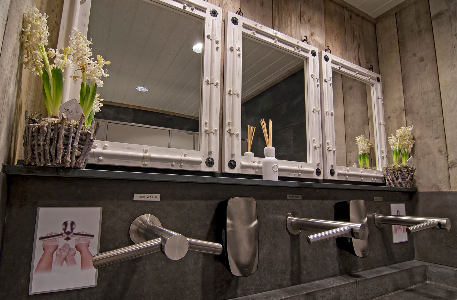 Badkamerwastafels met spiegels, handdrogers en bloemen. Rustiek hout en grijze oppervlakken. Instructies voor handen wassen zichtbaar.