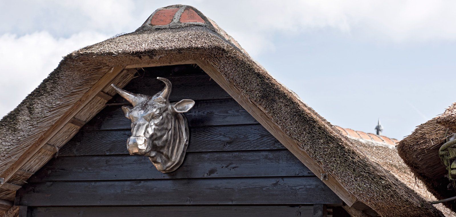 Een metalen stierenkop is gemonteerd op een houten gevel, met daarboven een rieten dak tegen een bewolkte hemel.