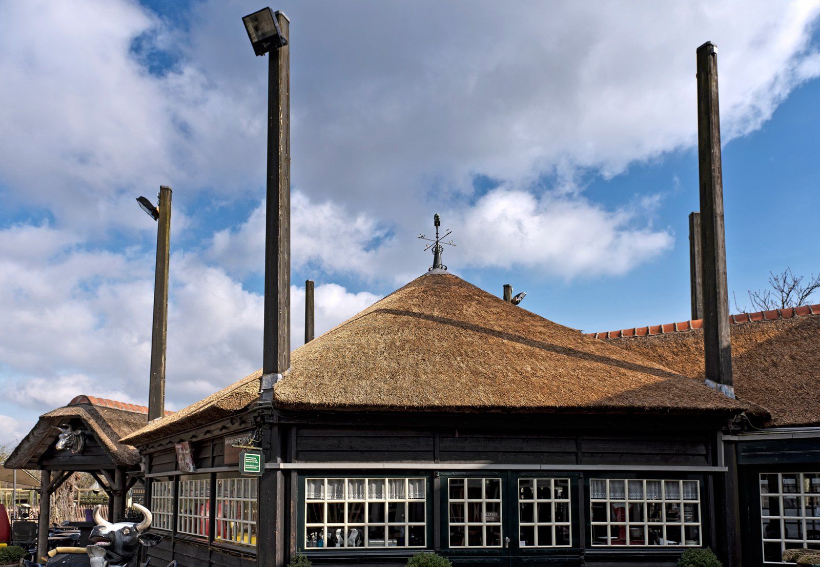 Een gebouw met een rieten dak, donkerhouten afwerking, witte ramen en hoge houten palen tegen een bewolkte hemel.