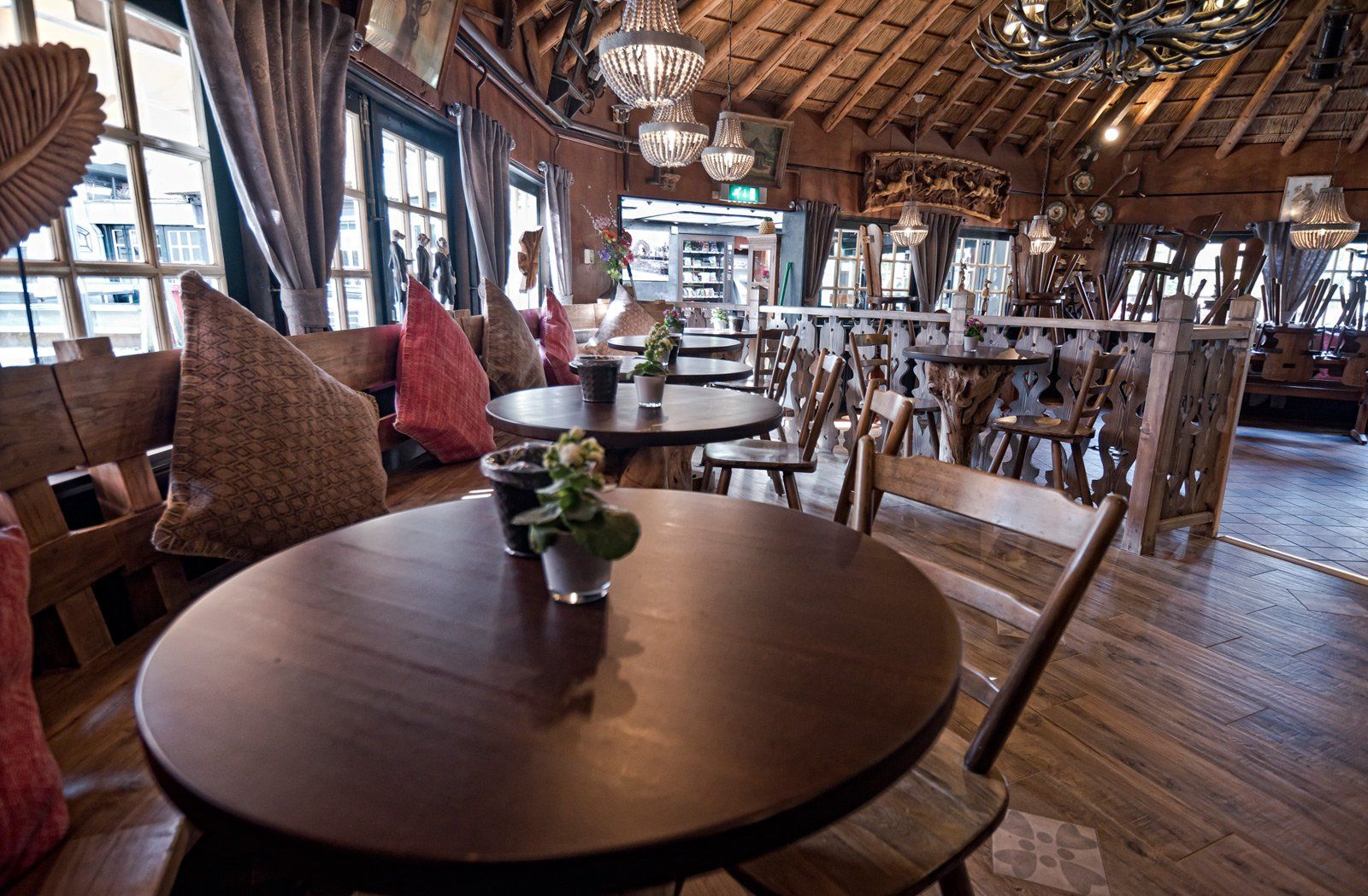 Restaurantinterieur met houten tafels, stoelen en een rustiek plafond. Ramen en decoratieve verlichting zijn zichtbaar.