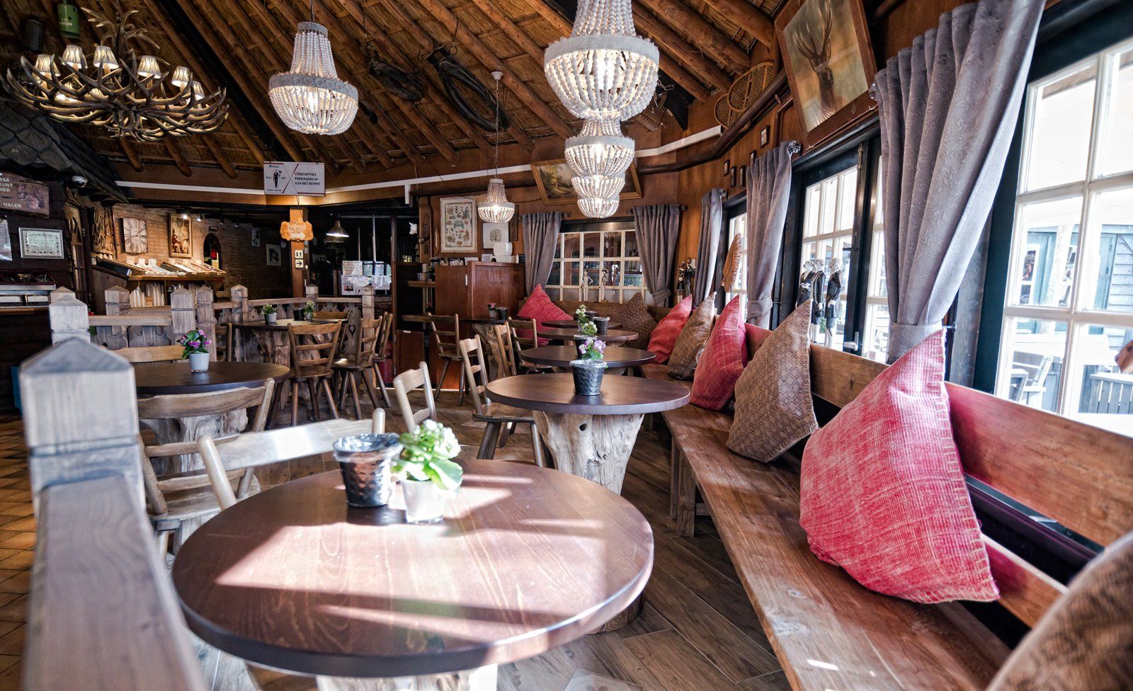 Gezellig restaurantinterieur met houten tafels, zitbanken en hangende kroonluchters. Zonlicht stroomt door de ramen naar binnen.