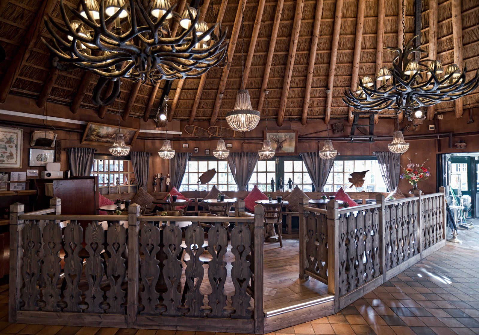 Restaurantinterieur met houtsnijwerk, kroonluchters, boogvensters en een rieten plafond.