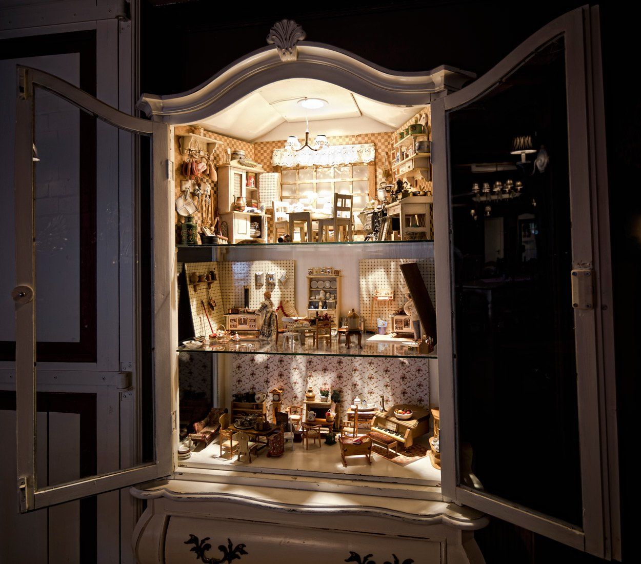 Een poppenhuis staat tentoongesteld in een witte kast, verlicht met miniatuurmeubels en -accessoires.