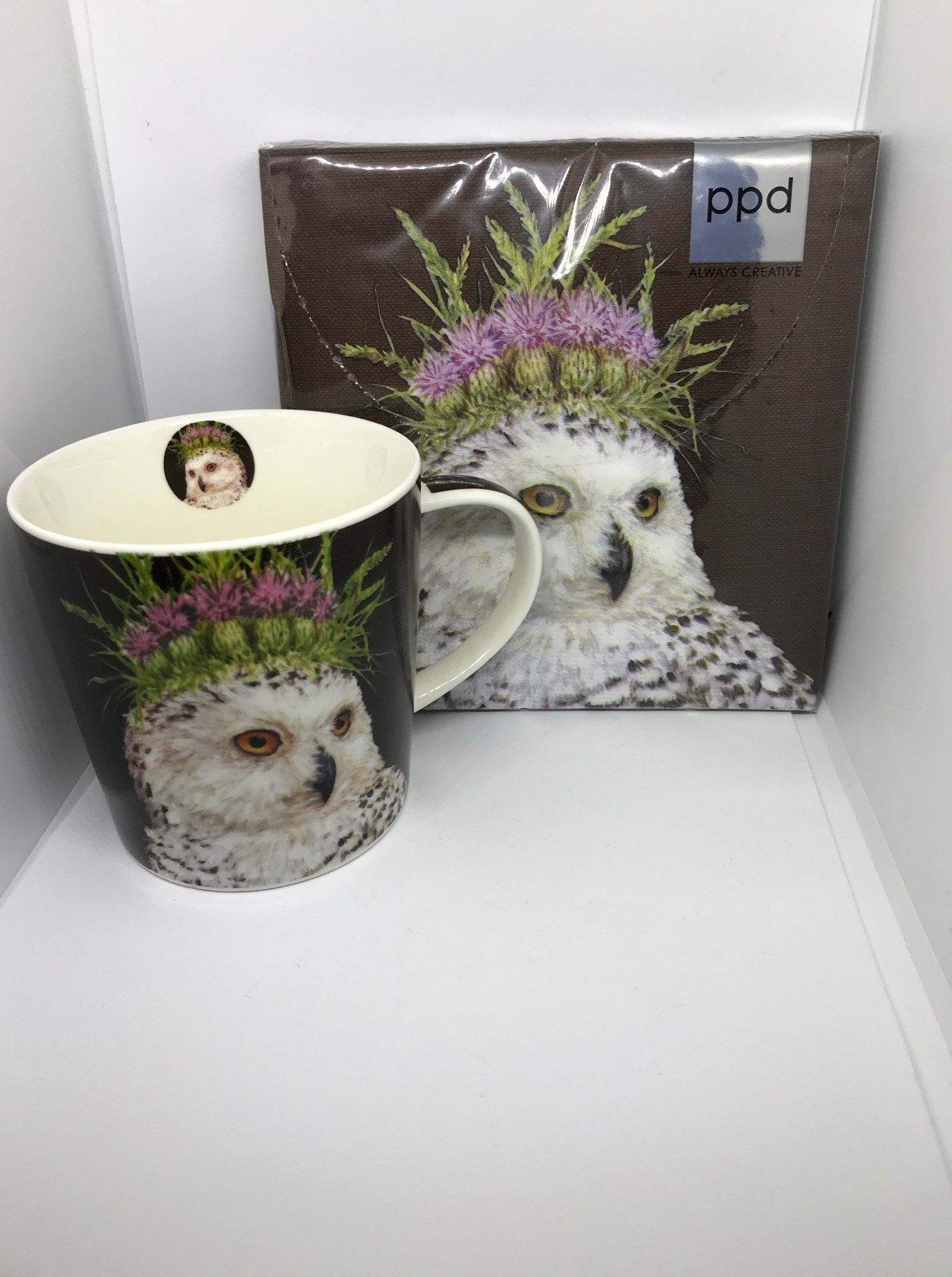 Mok en servet met een afbeelding van een uil met een bloemenkroon. De mok is zwart, het servet is bruin.