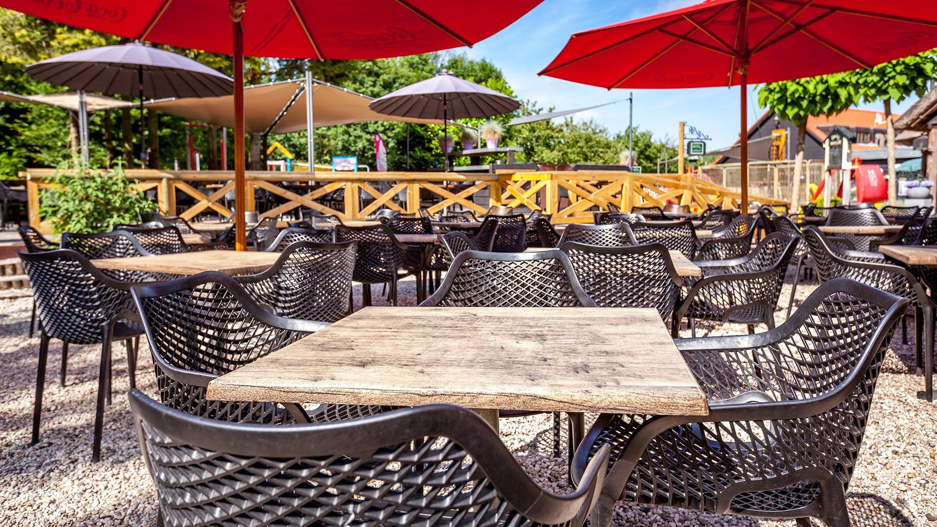 Terras van een restaurant met tafels, stoelen, rode parasols en een houten vlonder.