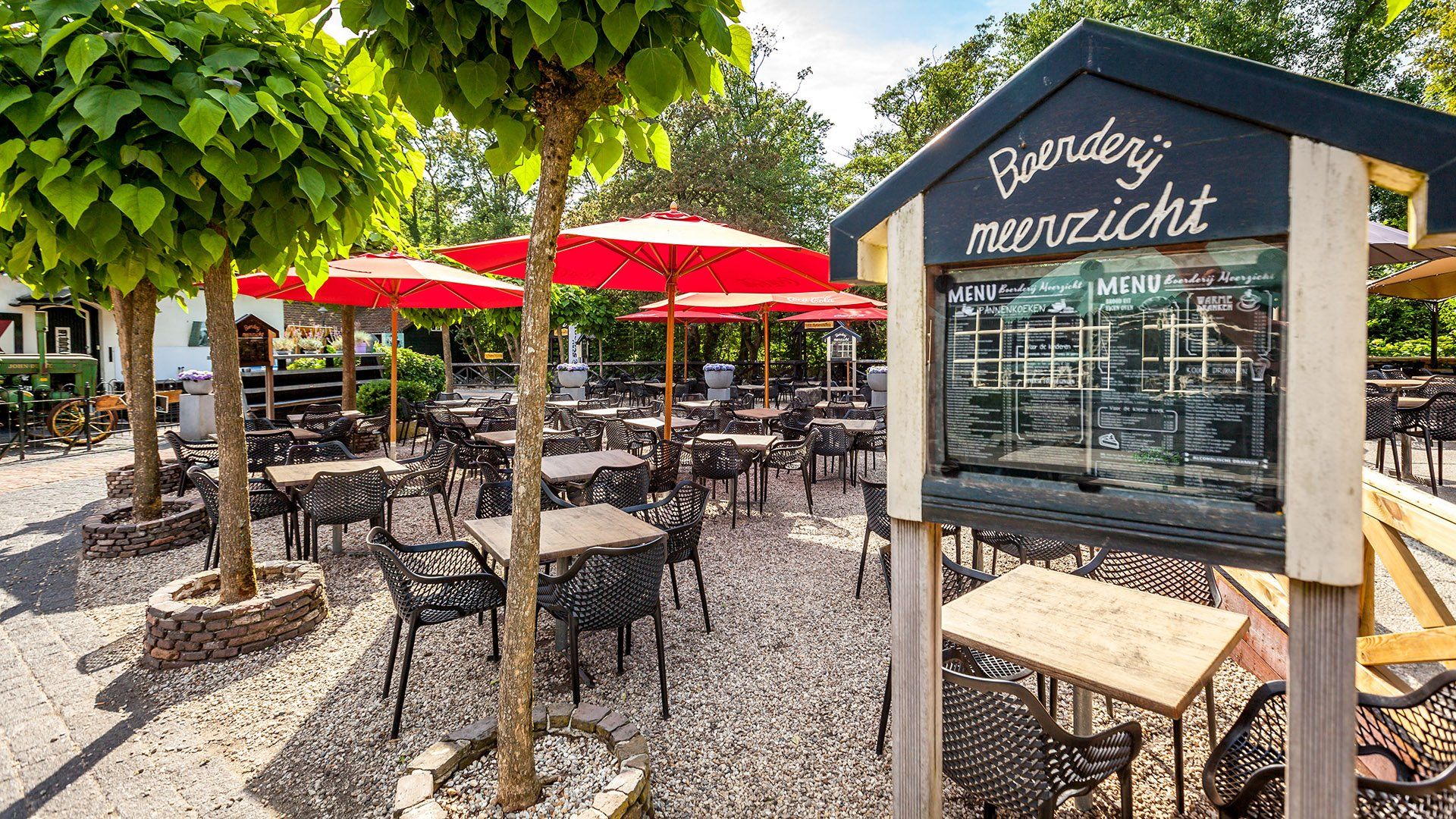 Een restaurant in de openlucht met tafels, rode parasols, bomen en een uithangbord.