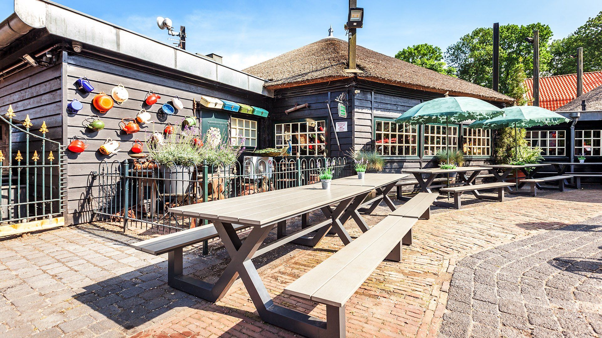 Buitenpatio met picknicktafels, kleurrijke potten aan de muur, parasols en een donkerhouten gebouw.