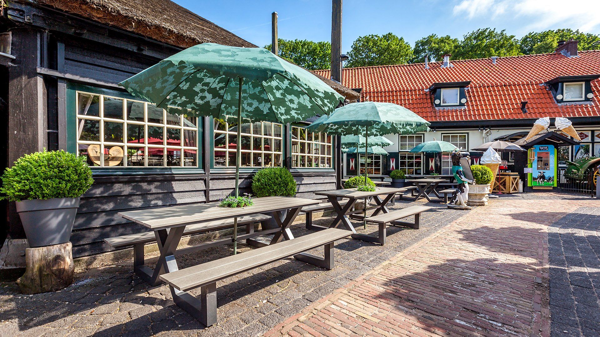 Buitenrestaurant met picknicktafels, groene parasols en een gebouw met een rieten dak.