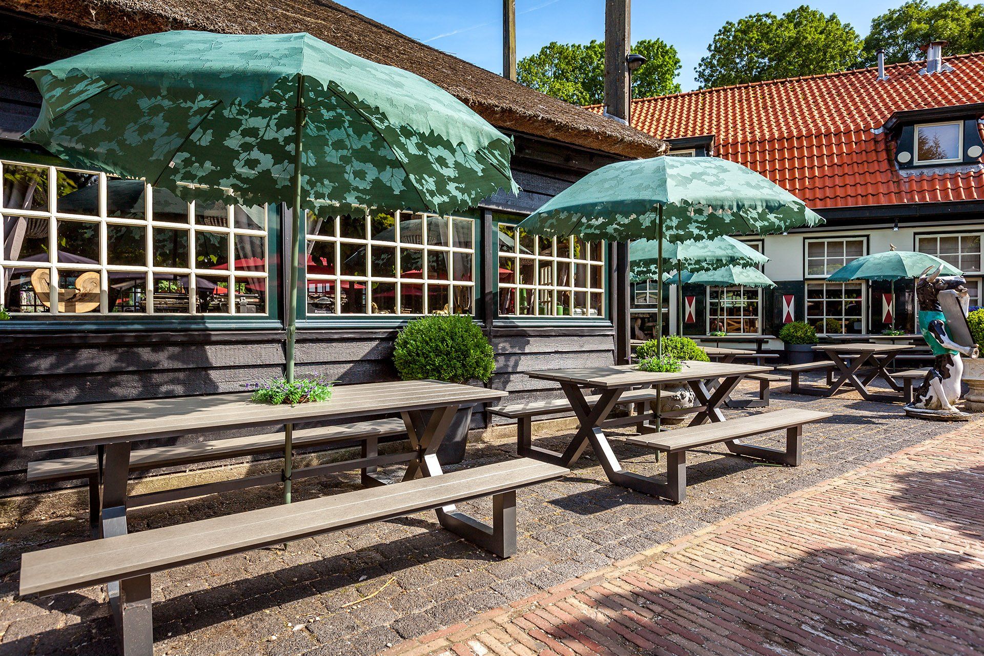 Een restaurant met terras, groene parasols, houten tafels en banken, en een gebouw met een rieten dak.