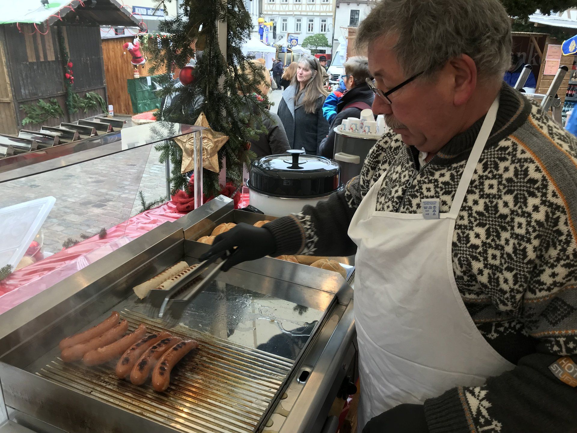 Ein Mann in einer Schürze brät Würstchen auf einem Grill.