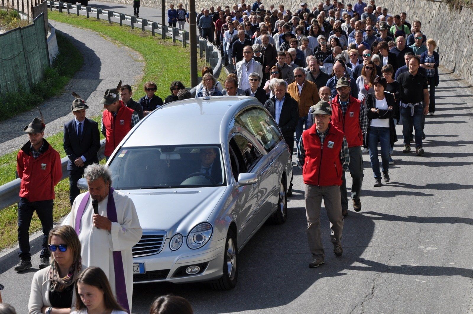 Un folto gruppo di persone sta camminando lungo una strada davanti a un carro funebre