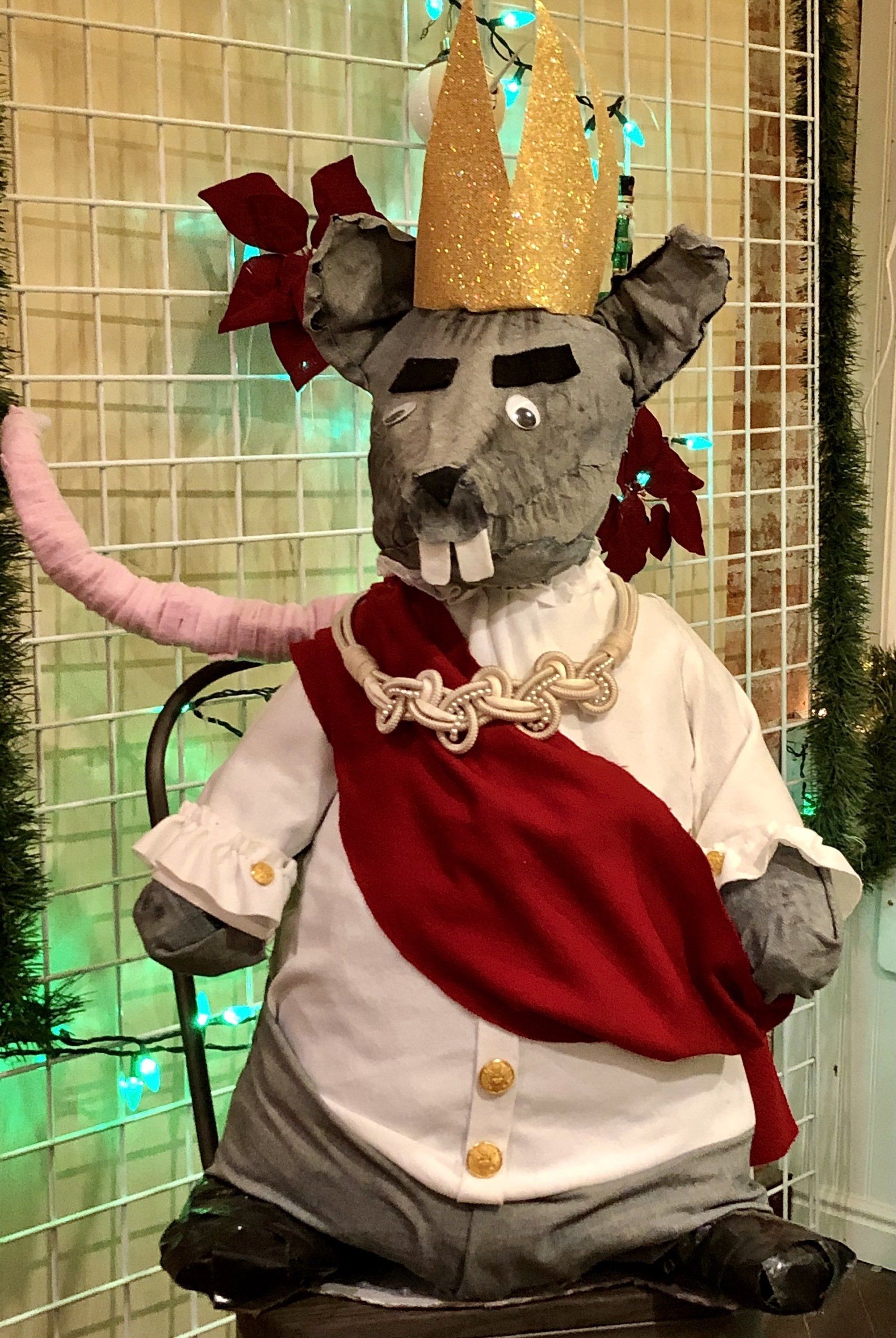 A stuffed mouse wearing a crown and a red sash is sitting on a chair.