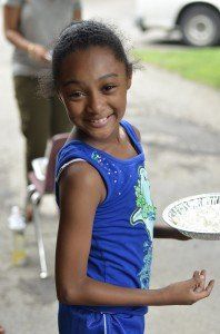 A young girl in a blue tank top is holding a plate and smiling.