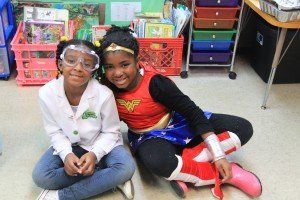 Two girls dressed in wonder woman costumes are sitting on the floor in a classroom.
