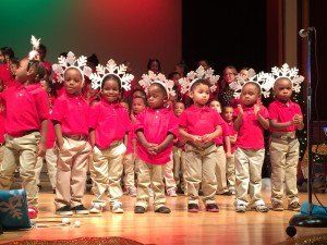 A group of children wearing red shirts and white pants are standing on a stage.