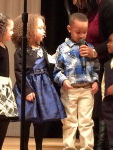 A boy is singing into a microphone while two girls look on