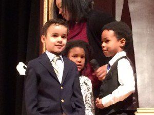A woman is holding a microphone in front of three children in suits
