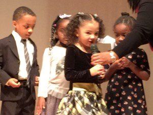 A group of children are standing in front of a woman holding a microphone