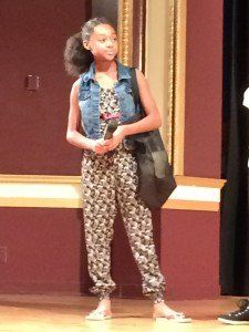 A young girl in a jumpsuit is standing on a stage