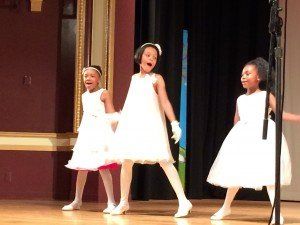 Three little girls in white dresses are dancing on a stage