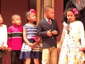 A group of children standing next to each other with one girl holding a microphone