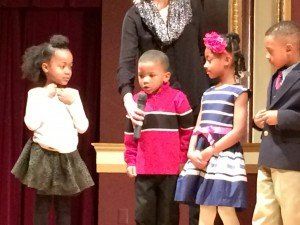 A group of children standing on a stage with a woman holding a microphone