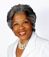 A woman with gray hair is wearing a white jacket and a pearl necklace.