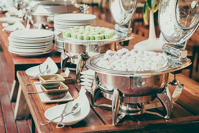 Een buffettafel met metalen warmhoudschalen gevuld met eten, stapels witte borden en serveerbestek.