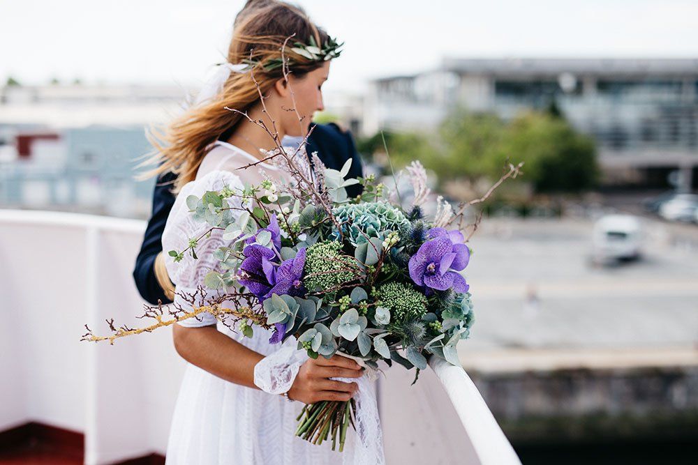 Een persoon in een witte jurk houdt een groot, kleurrijk boeket wilde bloemen vast terwijl ze op een terras staat met uitzicht op een haven.