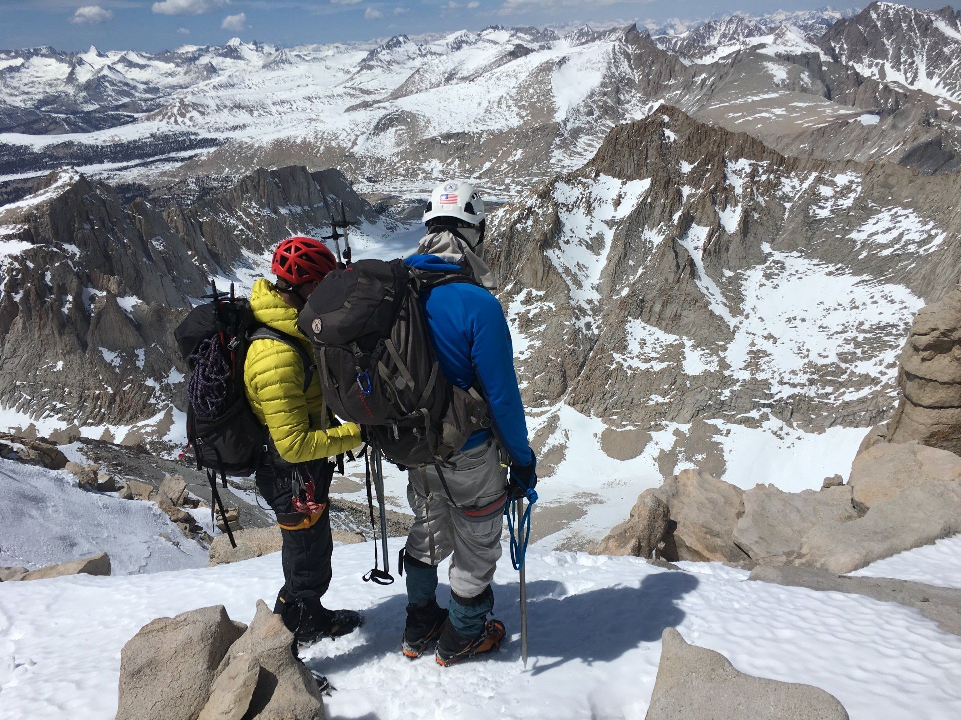 Two hikers in winter gear stand atop a snowy mountain, overlooking a rugged landscape.